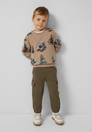 Młody chłopiec stojący przy szarej ścianie, ubrany w beżową bluzę z nadrukami pojazdów, oliwkowe spodnie bojówki i białe adidasy.