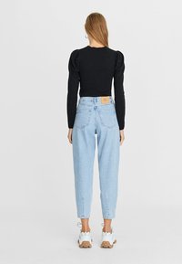 Frau mit glattem, hellbraunem Haar, die ein schwarzes Top mit Puffärmeln, hellblaue, hochgeschnittene Jeans und weiße Sneakers trägt, steht mit dem Rücken zur Kamera.