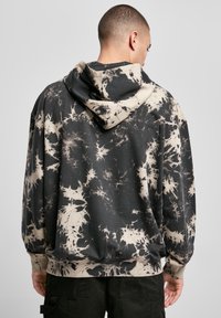 Bărbat îmbrăcat cu un hanorac negru și bej cu efect tie-dye, văzut din spate, asortat cu pantaloni negri, pe un fundal simplu de gri deschis.
