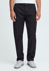Man in zwarte broek en grijze sneakers staat tegen een lichte achtergrond, met ontspannen handen aan zijn zijden. Casual stijl benadrukt.