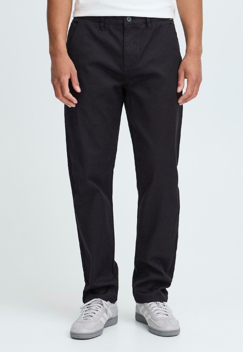 Man in zwarte broek en grijze sneakers staat tegen een lichte achtergrond, met ontspannen handen aan zijn zijden. Casual stijl benadrukt.