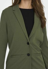 Zielona oliwkowa marynarka wykonana z gładkiego materiału, o dopasowanym kroju, z klapami wciętymi, dwoma przednimi kieszeniami i czarnymi guzikami.