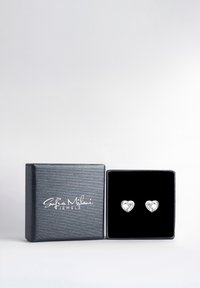 Silver hjärtformade örhängen visade i en svart sammetstapet box med en texturerad grå utsida och vit logotyp.