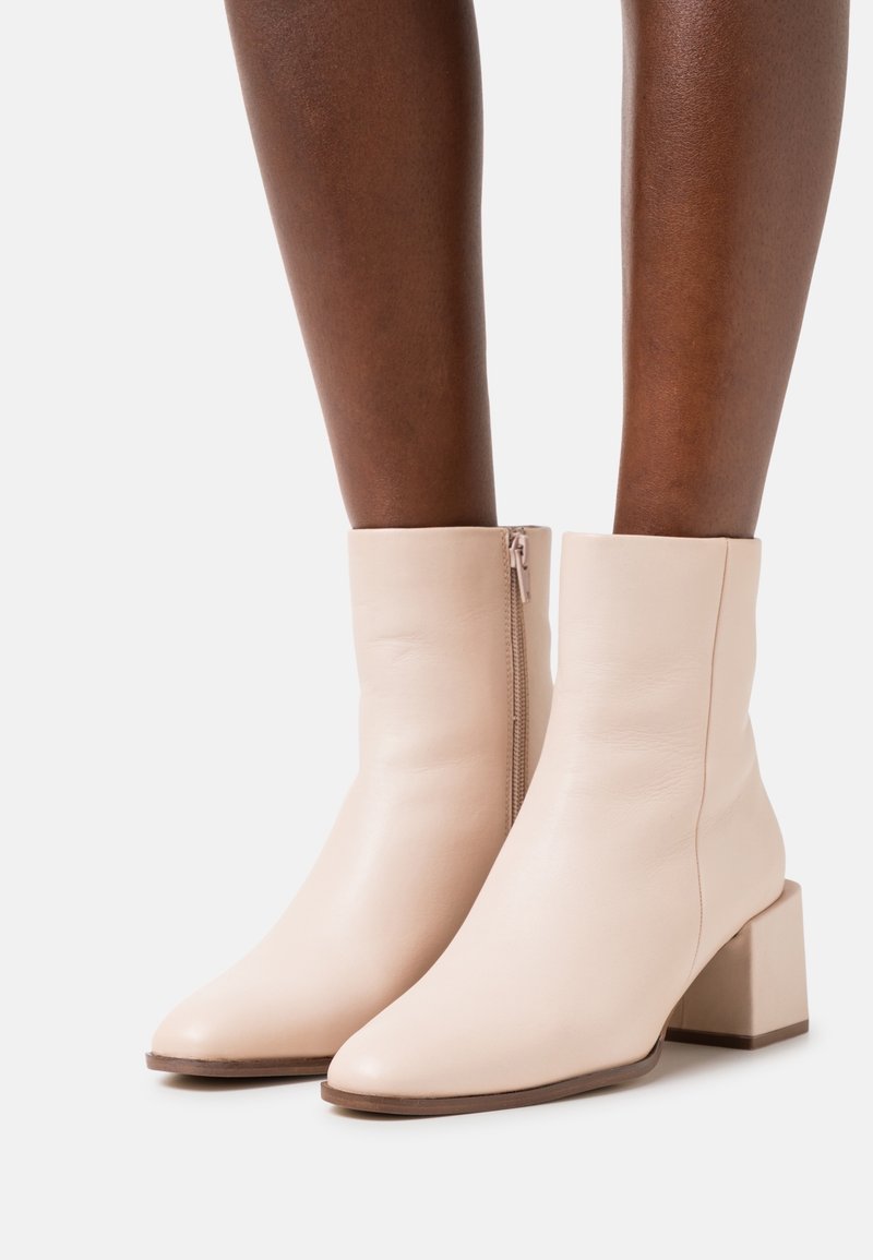 Bottes en cuir crème à hauteur de cheville avec talons carrés et fermetures éclair latérales, portées sur des jambes à peau foncée sur un fond uni.