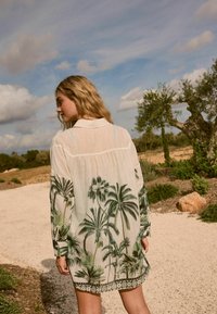 Doorzichtige witte shirtjurk met een groen palmblaadjesprint, lange mouwen, een kraag en een gegolfde onderzoom met zwarte accenten.