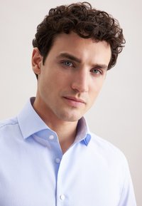 Lichtblauw gestreept shirt met een button-down kraag, gladde stof en een getailleerd ontwerp. Het model heeft krullend haar en kijkt naar de camera.