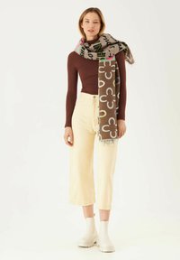 Bruine sjaal met patroon en franjes, versierd met kleurrijke bloemen en tekst, gedrapeerd over een model in een aansluitend long-sleeve top en een wijde broek.