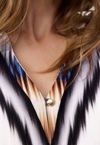 Mehrfarbige Bluse mit V-Ausschnitt, auffälligen grafischen Mustern in Schwarz, Weiß und Braun sowie einem metallischen Knopfdetail.