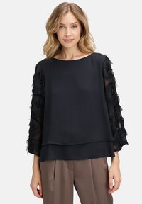 Blusa nera con design a strati, featuring maniche trasparenti decorate con frange nere. Tessuto liscio con scollatura rotonda e maniche ampie.