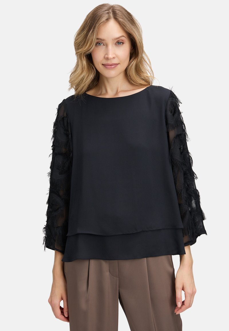 Blusa nera con design a strati, featuring maniche trasparenti decorate con frange nere. Tessuto liscio con scollatura rotonda e maniche ampie.