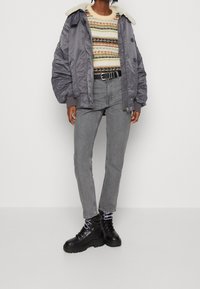 Chaqueta bomber gris acolchada con cuello de borrego, suéter de punto a rayas, pantalones vaqueros grises y botas negras con cordones y suela gruesa.