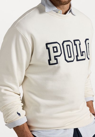 Kremni pulover s kapuco z vezenjem "POLO" v mornarsko modri barvi. Nosijo ga čez svetlo modro črtasto srajco s kratkimi rokavi. Ima logotip na rokavu.