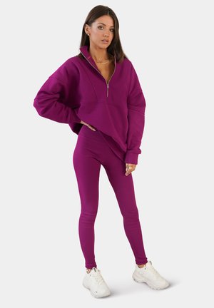 Lila Pullover mit Halbreißverschlusskragen und gesenkten Schultern, kombiniert mit passenden Leggings. Das Model trägt weiße Sneakers und präsentiert einen lässigen Schnitt.