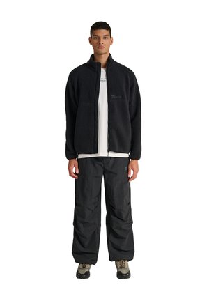 Młody mężczyzna stoi, ubrany w czarną polarową kurtkę na zamek, luźne czarne spodnie, białą koszulkę z grafiką oraz beżowe buty trekkingowe na białym tle.