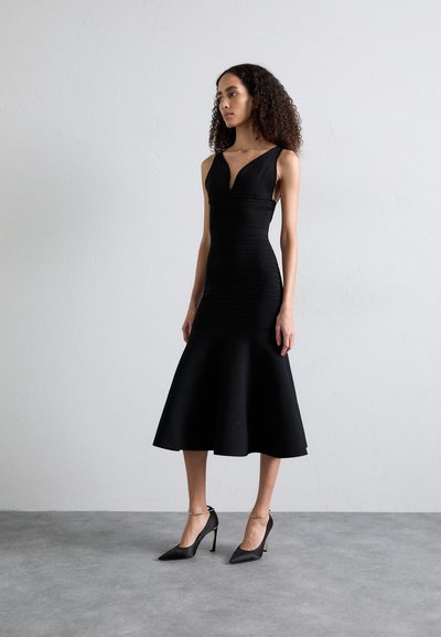Vestido negro ajustado con escote en forma de V y dobladillo acampanado. Textura suave, diseño sin mangas. Combinado con tacones negros de punta con correas en el tobillo.