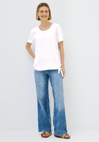 Camiseta blanca de manga corta con cuello redondo y detalle de nudo lateral, combinada con jeans acampanados de color azul claro y sandalias marrones.