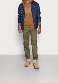 Giacca imbottita navy, felpa marrone con logo, pantaloni jogger verde oliva e sneakers beige con suola bianca. Design semplice, vestibilità casual.