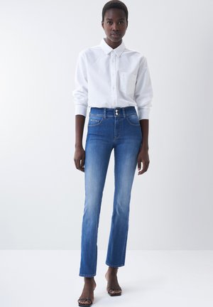 Jean en denim bleu taille haute avec un léger dégradé. Comprend deux poches avant, une fermeture à bouton et une coupe ajustée. Associé à une chemise blanche à boutons.