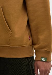 Καφέ φούτερ με καγκουρό τσέπη και ραμμένες μανσέτες, με πράσινη ετικέτα Barbour και μια μικρή κόκκινη ετικέτα Levi's στο πλάι.