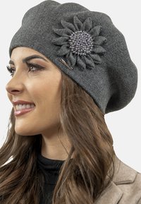 Szürke kötött beret virággal, amely gyöngyökkel díszített az oldalán. Sima textúra, puha anyag és laza fazon.