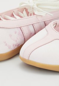 Sapatilhas atléticas rosas com parte superior em lona texturizada e couro liso, apresentando atacadores, sola de borracha e discretos bordados florais.