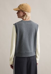 Graues ärmelloses Sweatshirt mit einem gerippten Ausschnitt, kombiniert mit einem hellgelben Langarmshirt darunter. Lässige Passform, dezente Struktur.