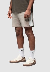 Shorts atletici beige con strisce laterali nere, abbinati a scarpe da ginnastica crema e marrone. Il modello indossa calzini bianchi e una maglietta verde scuro.