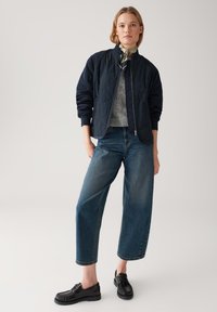 Navy gewatteerde bomberjack, grijze trui, wijde spijkerbroek in blauw, en zwarte veterschoenen, met een eenvoudige sjaal om de hals.