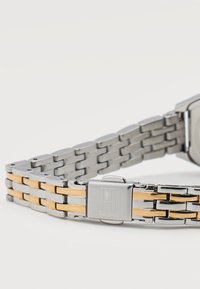 Βραχιόλι ρολογιού από ασημί και χρυσό μέταλλο με κούμπωμα που διαθέτει διακριτικό χαραγμένο λογότυπο, εμφανιζόμενο σε λευκό φόντο.