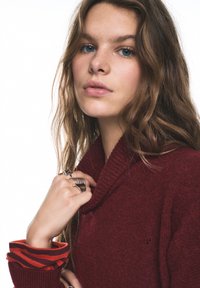 Viininpunainen neulepusero huivikauluksella, jossa on teksturoidu pinta ja kontrastiväriset raidalliset hihansuut punaisessa ja tummanpunaisessa.