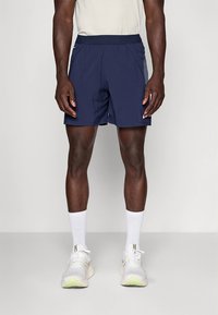 Calções atléticos azul-marinho feitos de tecido leve, com cintura elástica e acentos em painéis laterais. Usados com meias brancas e ténis.