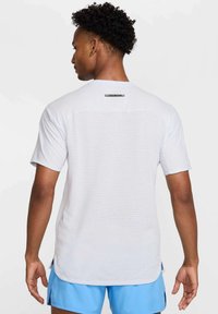 Camiseta deportiva de manga corta en un gris claro, con panel trasero texturizado y aberturas laterales para ventilación. Logotipo negro cerca del cuello.