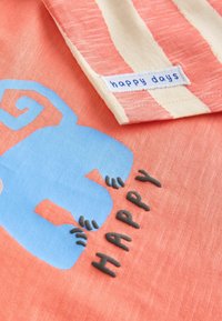 Korallenstoff mit einem grafischen Motiv eines blauen Affen und texturiertem Druck, der "HAPPY" buchstabiert. Enthält ein label mit gestreiftem cremefarbenen und pfirsichfarbenem Kragen.