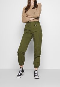 Oliwkowe spodnie cargo z elastycznymi mankietami, z ozdobnym kółkiem na talii, w połączeniu z beżową bluzką z długim rękawem i czarnymi trampkami.