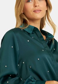 Turquoise satijnen blouse met een puntkraag, versierd met verspreide rhinestone accenten, en lange mouwen met gegatherde manchetten.