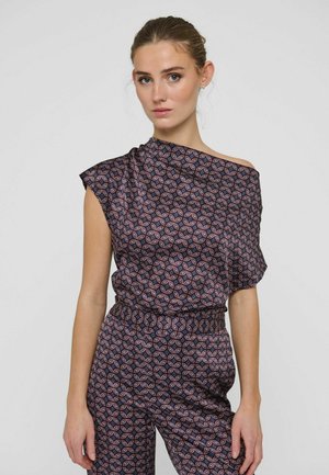 Donna che indossa un top monospalla scuro con motivo e pantaloni coordinati con un motivo geometrico rosa e blu, in piedi su uno sfondo semplice.