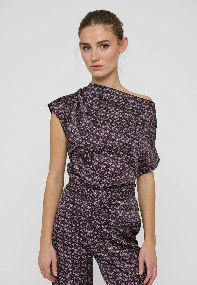 Femme portant un top asymétrique sombre à motifs et un pantalon assorti avec un motif géométrique rose et bleu, debout devant un fond uni.