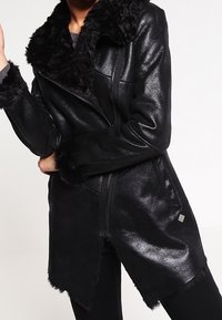 Schwarze Kunstlederjacke mit einem strukturierten Pelzkragen und Ärmelbündchen, ausgestattet mit einem Reißverschluss und einem asymmetrischen Saum.