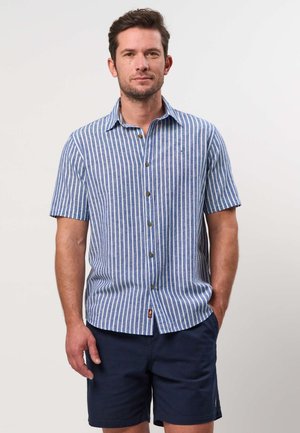 Hombre con camisa de botones de manga corta a rayas verticales azules y blancas y pantalones cortos azul marino, de pie sobre un fondo claro y liso.