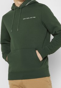 Dark green hoodie made of soft fabric, featuring a kangaroo pocket, drawstring hood, and white text: "NEW YORK, EST. 1985."