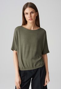 Top verde oliva a maniche corte con tessuto testurizzato, scollatura rotonda e orlo arricciato. Abbinato a pantaloni neri.