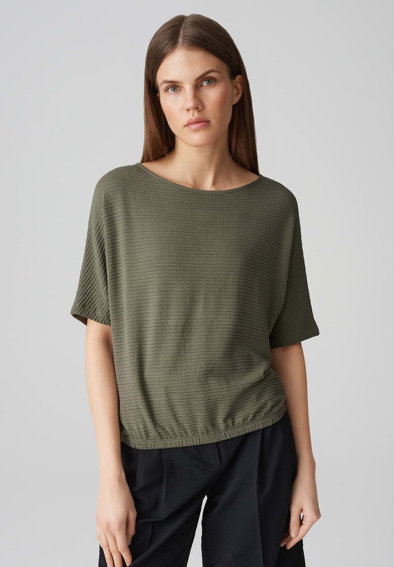 Top verde oliva a maniche corte con tessuto testurizzato, scollatura rotonda e orlo arricciato. Abbinato a pantaloni neri.