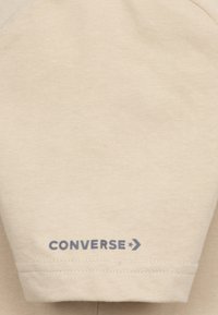 Beige színű pamut pólóujj, amelyen nyomtatott kék "CONVERSE" logó és nyíl díszítés található, sima textúrával és egyszerű dizájnnal.