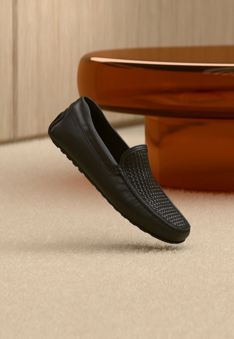 Mocassin en cuir noir avec un détail tissé sur le dessus flottant au-dessus d'un tapis beige dans un intérieur moderne avec une table ronde marron.