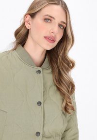Jachetă bomber matlasată de culoare verde măslinie cu guler elastic, având butoane negre și un model în valuri pe țesătură.
