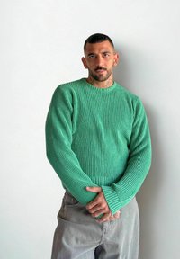 Mężczyzna z krótkimi włosami i tatuażami, ubrany w zielony sweter z prążkami i szare spodnie, stoi z założonymi rękami przy gładkiej ścianie.
