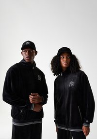 Vestes en velours noir avec fermeture éclair à l'avant, rayures blanches accentuées aux poignets et à l'ourlet, et logo brodé. Des casquettes noires complètent les tenues.