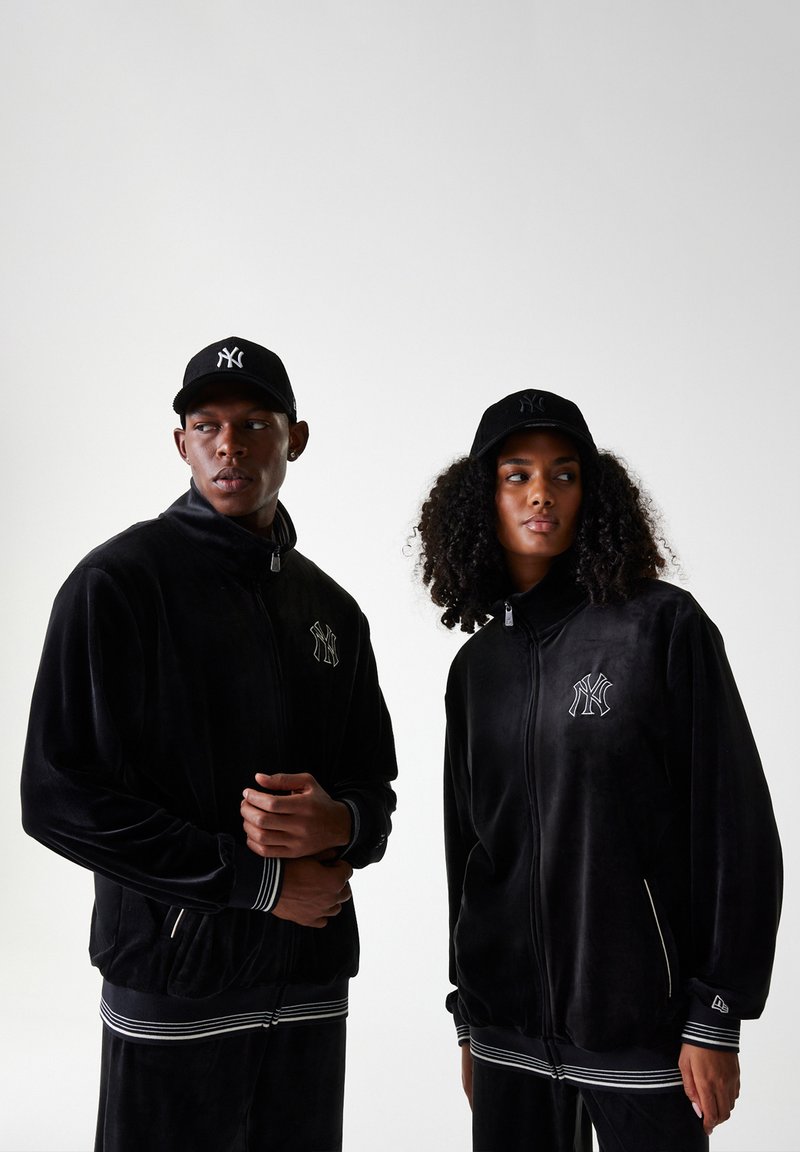 Vestes en velours noir avec fermeture éclair à l'avant, rayures blanches accentuées aux poignets et à l'ourlet, et logo brodé. Des casquettes noires complètent les tenues.