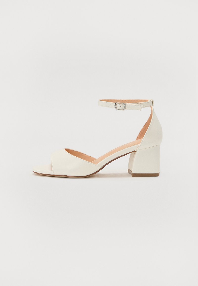 Lys cremefarvet hælesandal med ankelrem, åben tå og blokhæl. Glat lædertekstur med minimalistisk design.