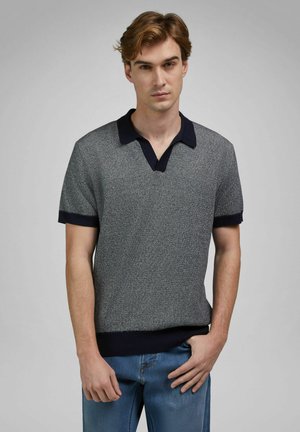 Giovane uomo con capelli castano chiaro che indossa una polo a maniche corte in maglia grigia con colletto e polsini blu navy scuro, abbinata a jeans blu, in piedi davanti a uno sfondo grigio.